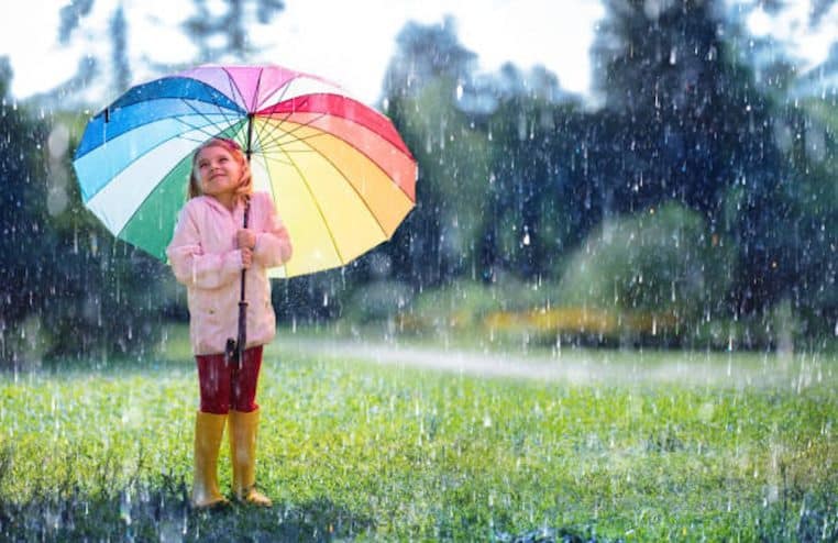Petite fille avec un parapluie multicolore qui sourit sous les averses dans un jardin.