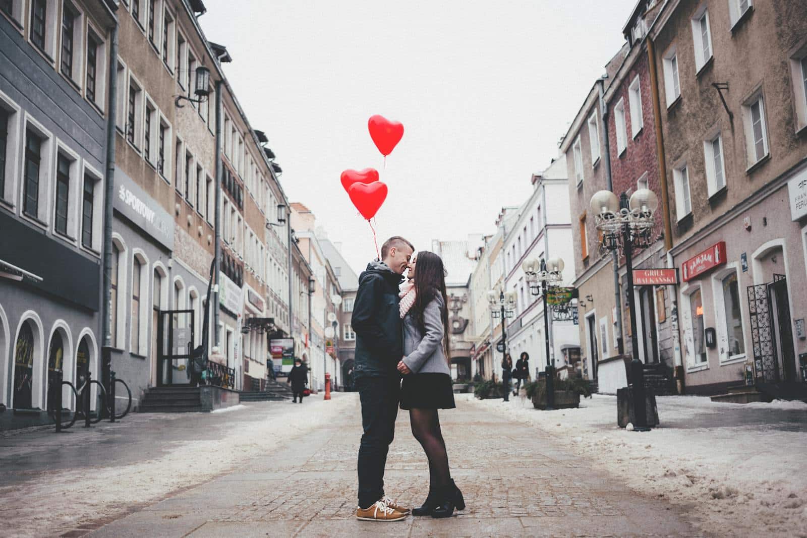A loving couple shares a kiss with heart-shaped balloons in a charming urban street setting.