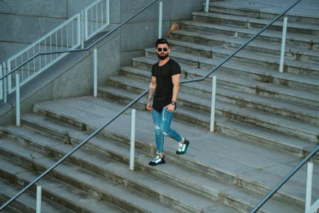 Casual bearded man in denim strolling down city steps with sunglasses and tattoos.