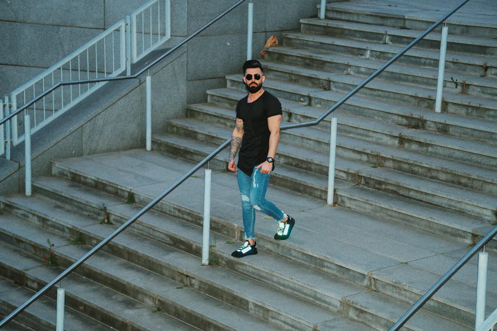 Casual bearded man in denim strolling down city steps with sunglasses and tattoos.