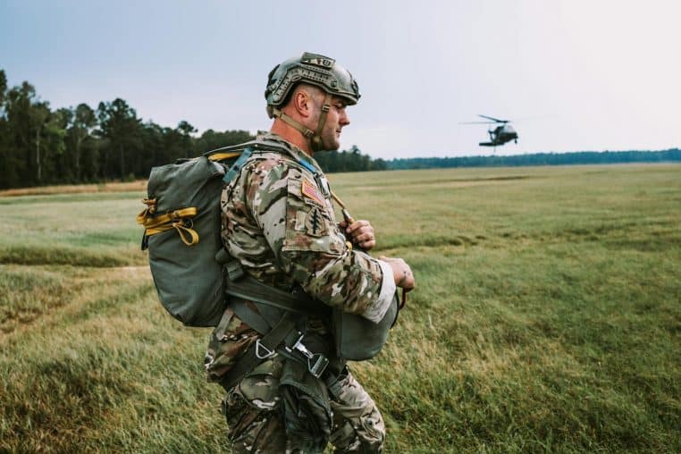 A soldier in camouflage walking in a field with a helicopter overhead.