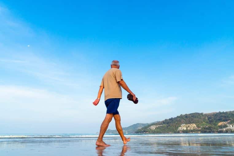 Homme âgé marchant pieds nus sur une plage, mer à l’arrière-plan