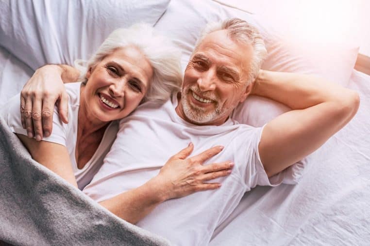 Deux seniors souriants allongés dans un lit sous une couette claire.