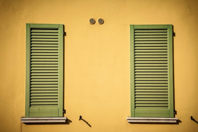 Two green shuttered windows on a vibrant yellow wall, captured in daylight.