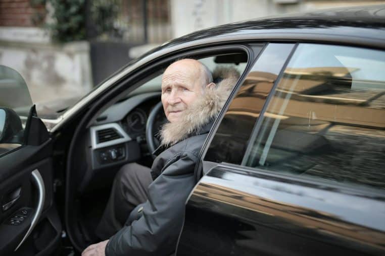 An elderly man in a winter coat stepping out of a parked car outdoors.