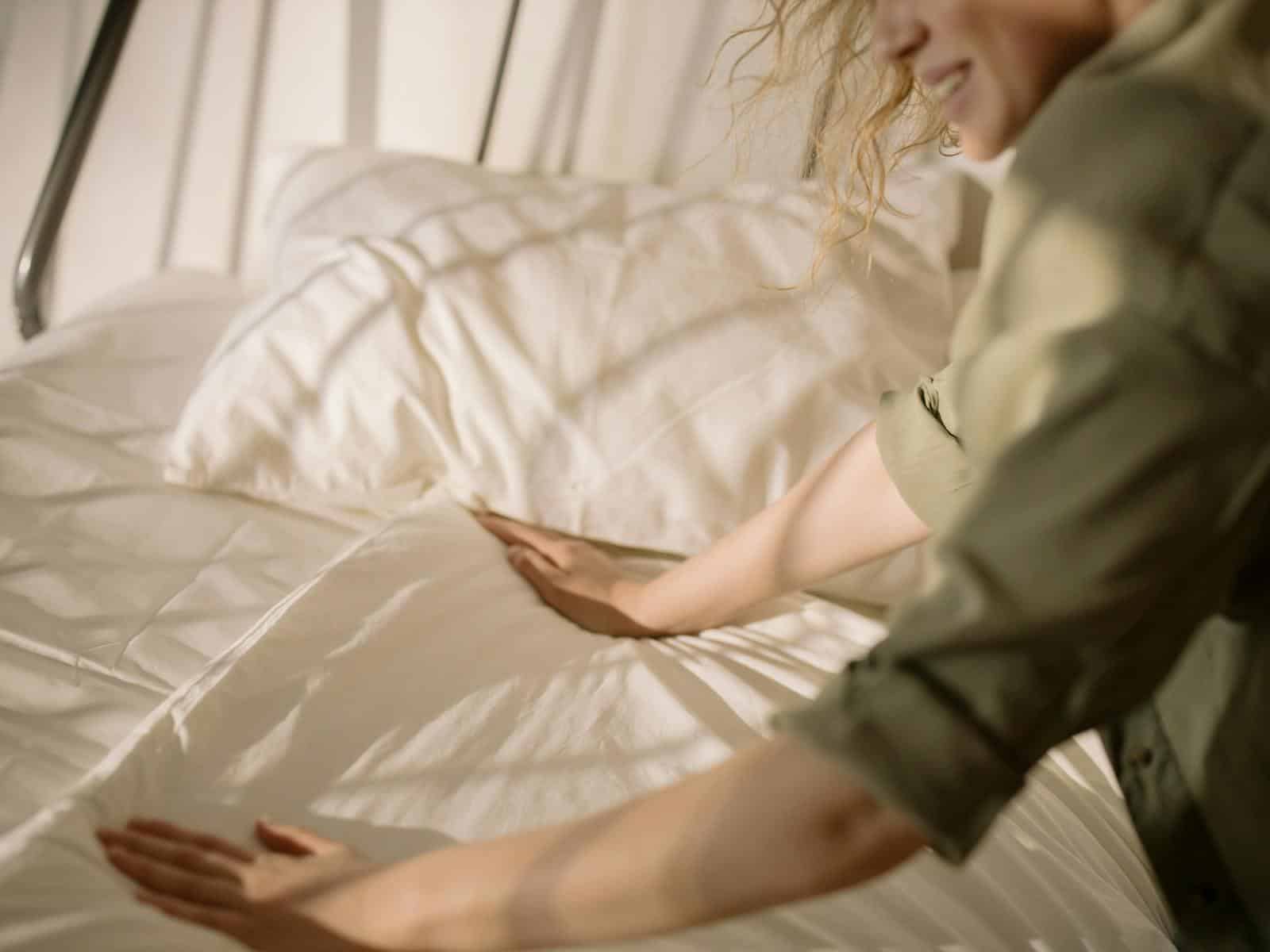 A person arranging bed linens in a warm, sunlit bedroom, creating a cozy and inviting atmosphere.