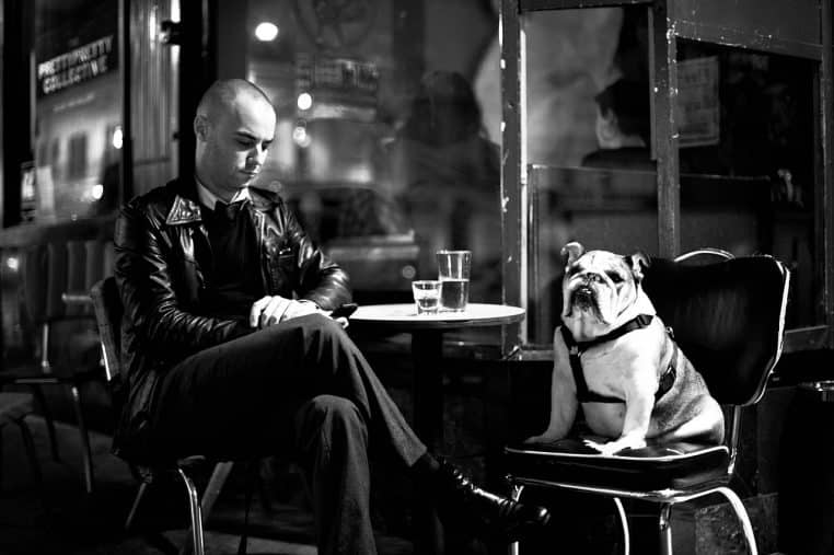 Homme en veste en cuir assis à une table de café, smartphone en main, un bouledogue assis sur la chaise voisine.