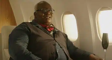Magloire assis dans un siège d’avion, portant des lunettes et un foulard rouge, lors d’une interview.