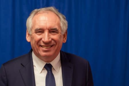 François Bayrou souriant, costume bleu, devant un rideau bleu prolongé sur toute la largeur.