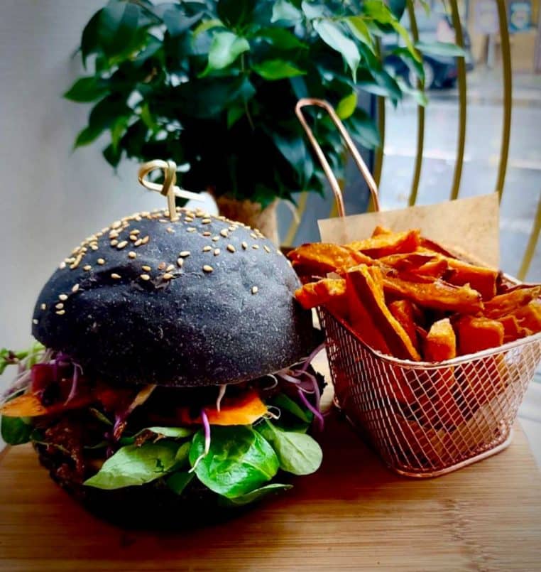 Burger au bun noir garni de jeunes pousses et légumes, frites de patate douce en corbeille.