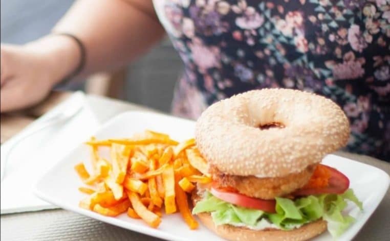 Bagel au pain sésame, poulet croustillant, tomate, laitue et frites de patate douce sur assiette blanche.