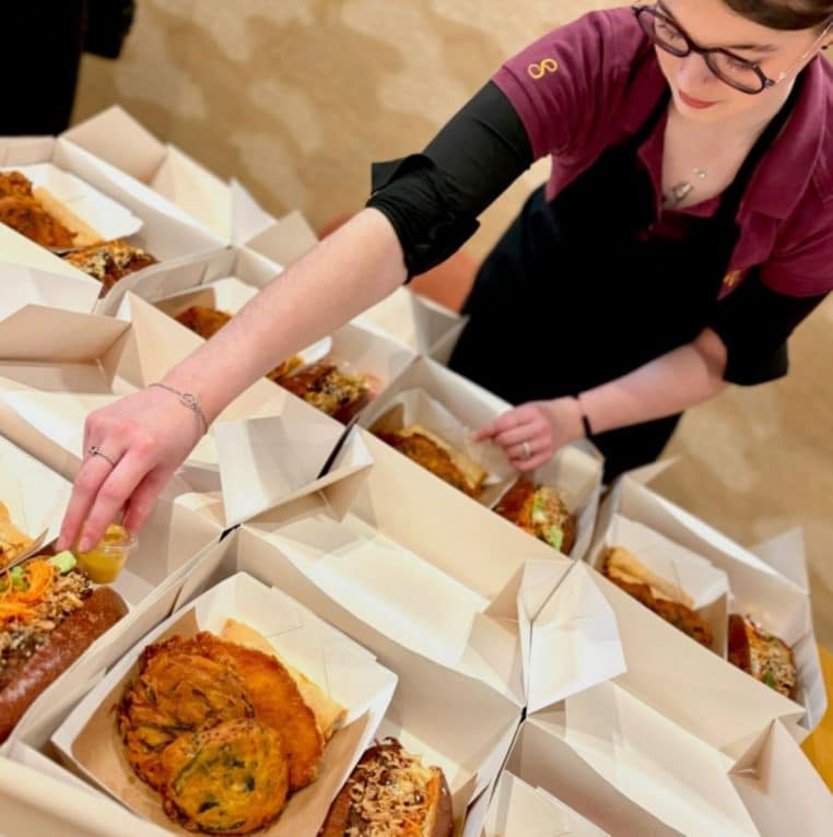 Employée qui garnit des box repas avec assortiments croustillants et condiments.