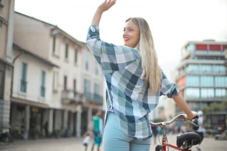Blonde woman waving while holding a bicycle in a charming Italian street setting.