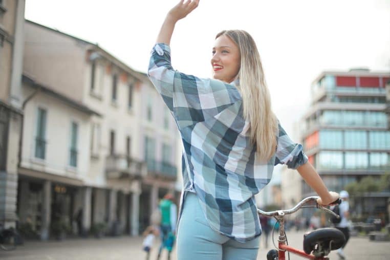 Blonde woman waving while holding a bicycle in a charming Italian street setting.