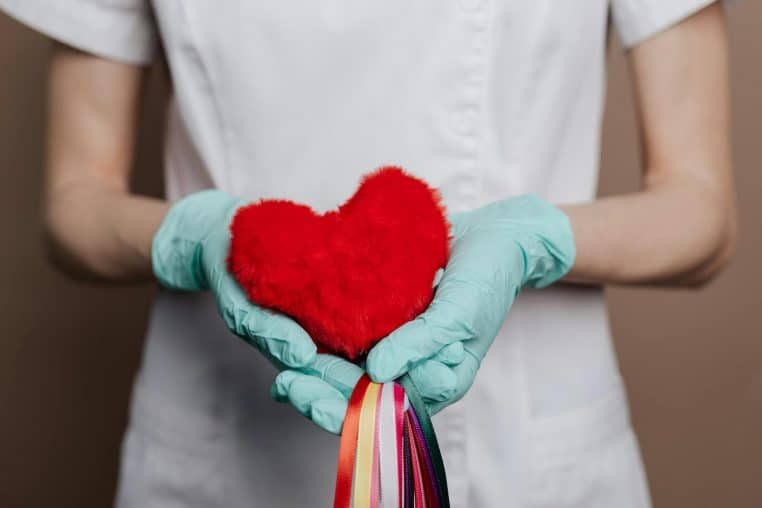 A nurse in gloves holds a red heart with rainbow strings, symbolizing love and healthcare.