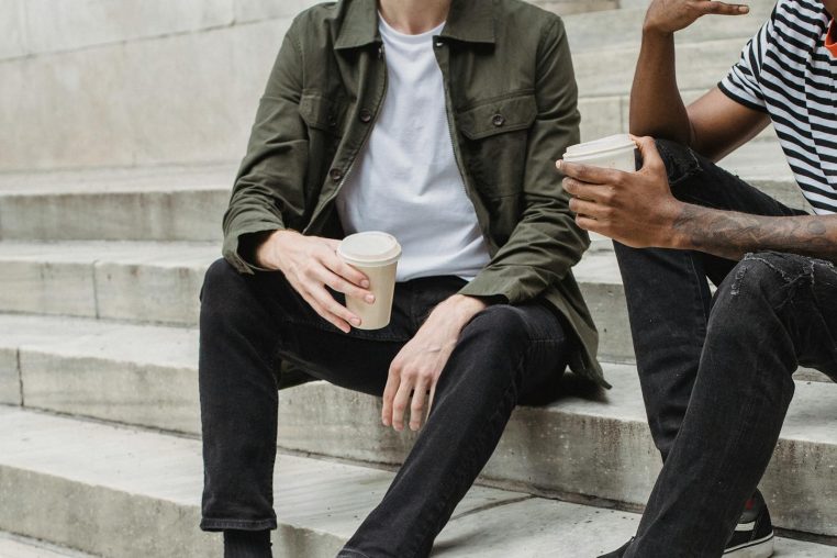 Unrecognizable multiracial male with paper cups of hot drinks sitting on concrete stairs of university building on street during coffee break