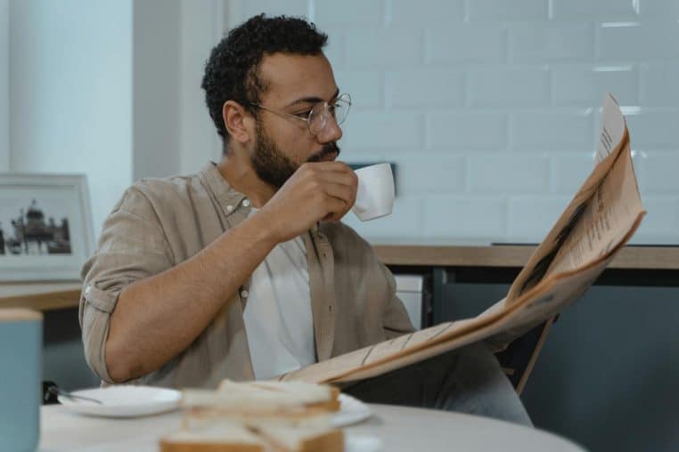 Bearded man with glasses reads newspaper while sipping coffee in a modern kitchen.