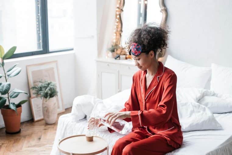 A woman in red pajamas pours water from a carafe into a glass, sitting on a bed in a cozy bedroom.