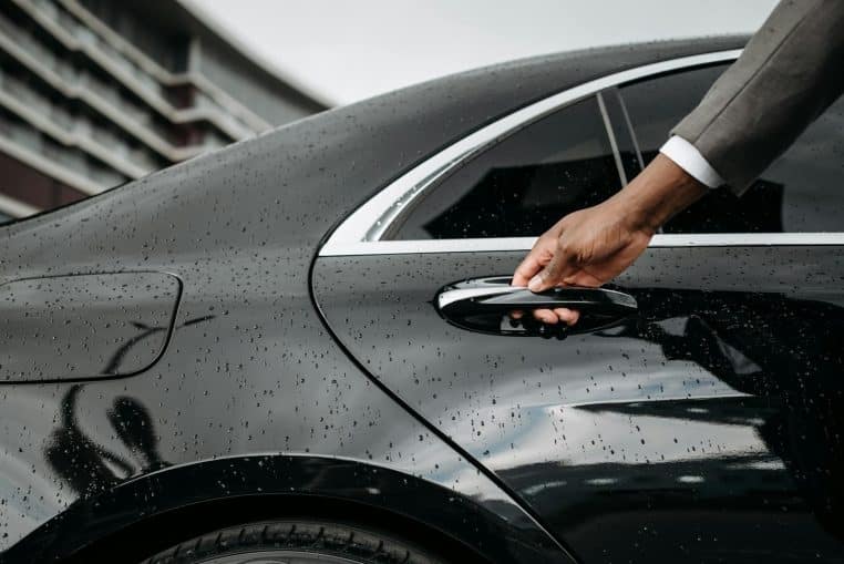 A close-up of a hand opening a black car door covered in raindrops, showcasing elegance and weather detail.