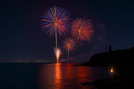 Bouquets de feu d’artifice au-dessus de la mer, reflets rouges et bleus sur l’eau, silhouette d’un phare en arrière-plan lors d’un hommage nocturne.