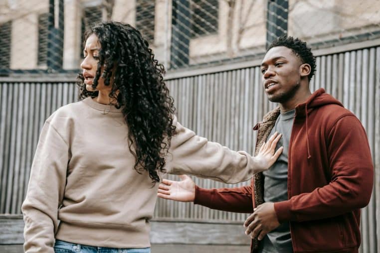 A couple in casual clothing appears to be in a disagreement outdoors with grey fencing background.