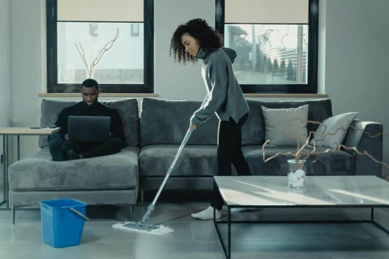 Man working on laptop while woman mops floor in stylish living room.