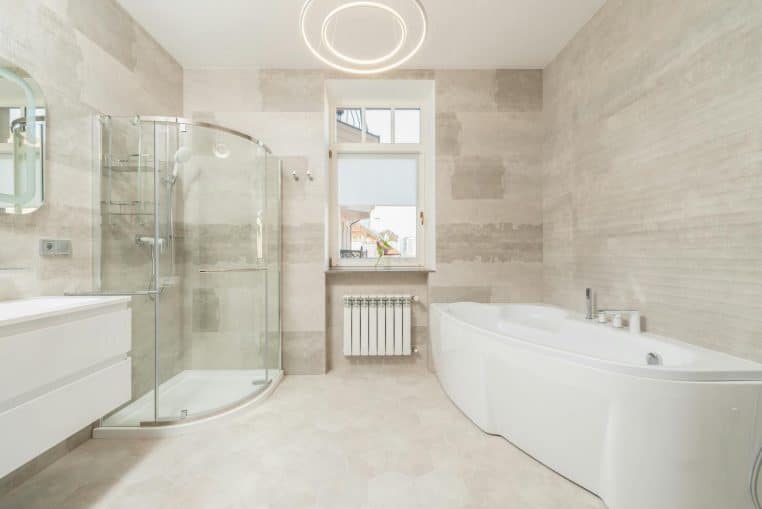 Modern spacious light bathroom with white bath and glass shower cabin near window