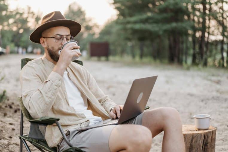 Man enjoying coffee and laptop work in nature, showcasing digital nomad lifestyle.
