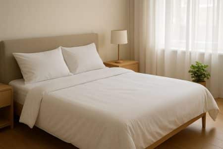Lit double fraîchement fait avec draps blancs dans une chambre lumineuse, rideaux voilés et plante verte sur parquet clair.