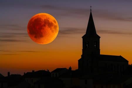 Lune de sang rouge cuivré se levant au-dessus d’un clocher au crépuscule, ciel orangé et fins nuages lors de l’éclipse du 7 septembre 2025.