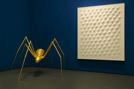 Sculpture d’araignée en métal doré face à une grande toile blanche en relief, accrochée sur un mur bleu nuit dans une galerie d’art.