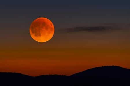 Lune de sang au lever, rouge cuivré, au-dessus d’un horizon est dégagé au crépuscule, avec silhouettes de collines en contre-jour.
