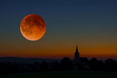 Éclipse lunaire en France : disque rougeoyant de la Lune de sang se levant au-dessus d’un clocher de village, ciel crépusculaire dégagé.