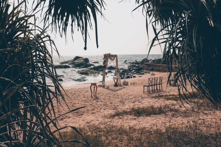 Arche florale et quelques chaises installées pour une cérémonie intime sur une plage encadrée de palmes, face à la mer et aux rochers.