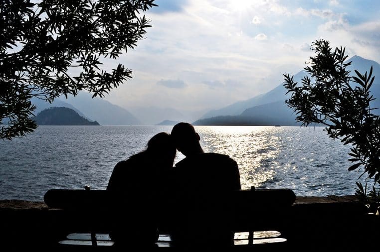 Silhouettes d’un couple assis face au lac au coucher du soleil, montagnes en arrière-plan et reflets dorés sur l’eau.