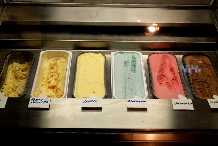 Rangée de bacs de glaces en vitrine réfrigérée (vanille, menthe, fruits, chocolat), étiquetés et alignés derrière la vitre.