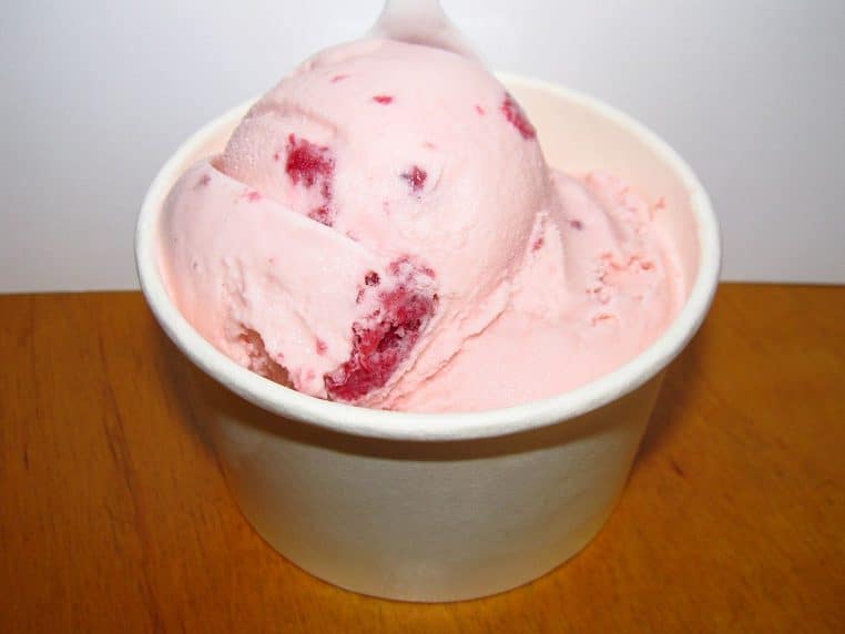 Pot blanc contenant une boule de glace à la fraise avec inclusions de fruits rouges, posé sur une table en bois clair.