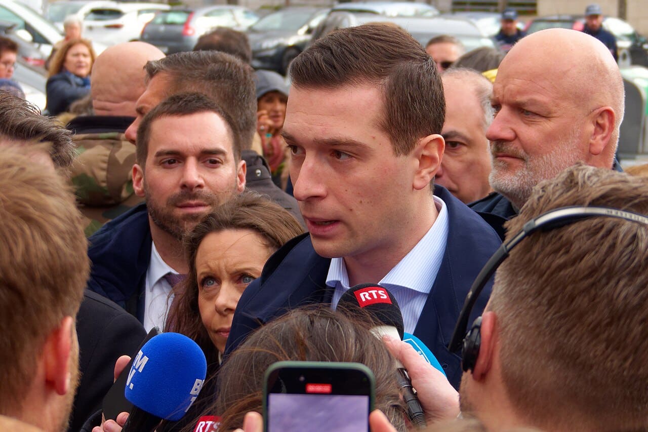 Jordan Bardella entouré de journalistes à Belfort, 23 mars 2024, répondant aux micros lors d’un déplacement de campagne.