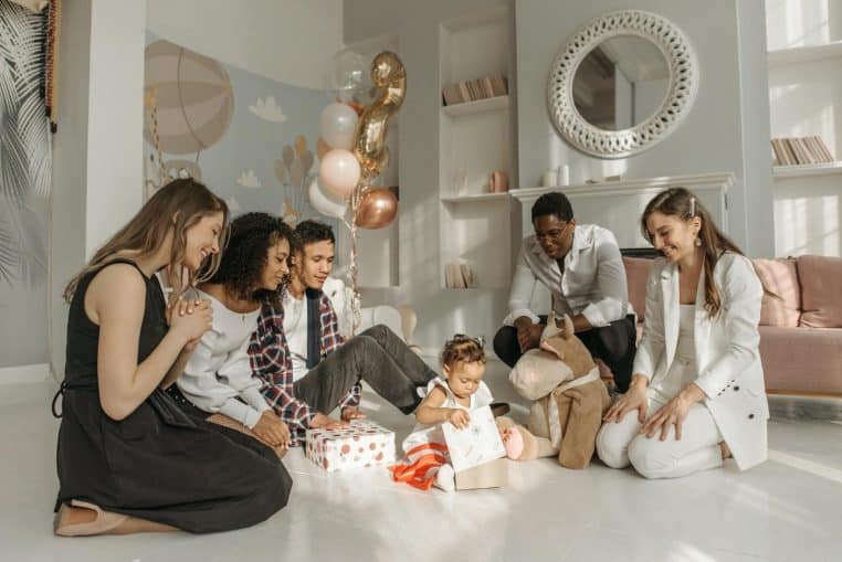 A diverse family celebrating a birthday with a baby girl opening gifts in a bright, festive room.