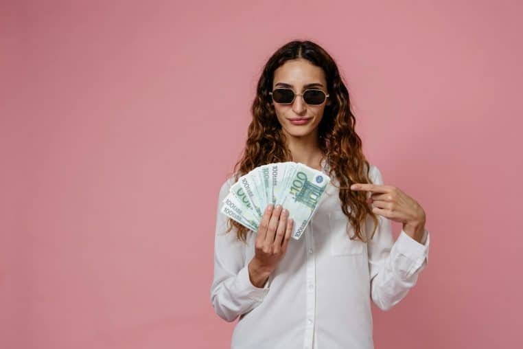 Woman in sunglasses with euro banknotes, expressing confidence and style against a pink backdrop.