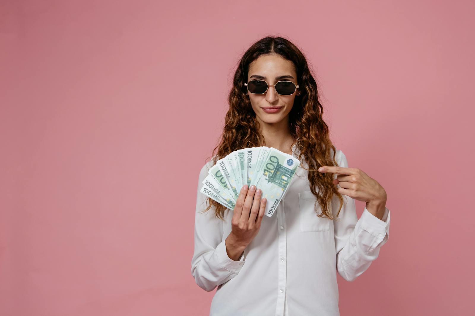 Woman in sunglasses with euro banknotes, expressing confidence and style against a pink backdrop.