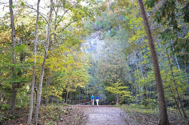 Un couple marche à l’automne sur un sentier tapissé de feuilles, falaises et forêt en bord de chemin.