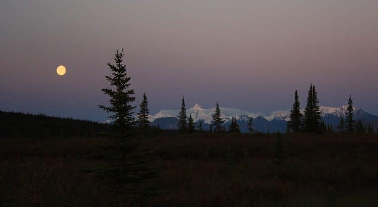 Pleine Lune blanche s’élevant sur la chaîne de l’Alaska, montagnes sombres et horizon dégagé, cadrage horizontal panoramique