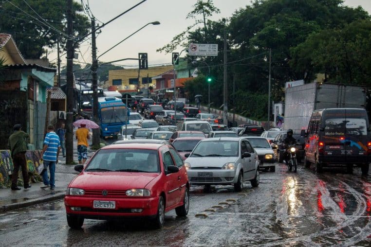 Trafic dense sur une artère de Taboão da Serra par temps de pluie, piétons et voitures partagent une chaussée saturée.