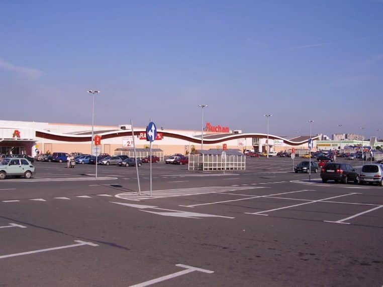 Vue lointaine d’un hypermarché Auchan avec vaste parking presque vide, montrant l’échelle des formats historiques