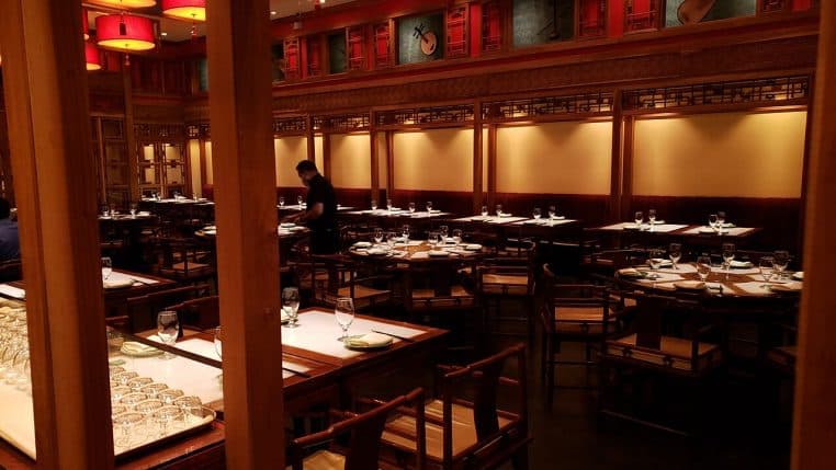 Salle de restaurant contemporaine en boiseries, tables carrées dressées et suspensions rouges, serveur préparant le service.