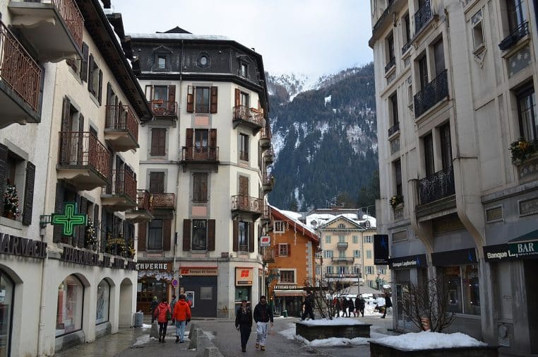 Passants dans une rue de Chamonix bordée d’immeubles alpins, montagne enneigée en toile de fond.
