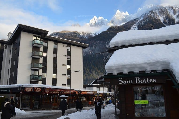 Place animée de Chamonix sous la neige, façades alpines et versant boisé avec pistes visibles au loin.