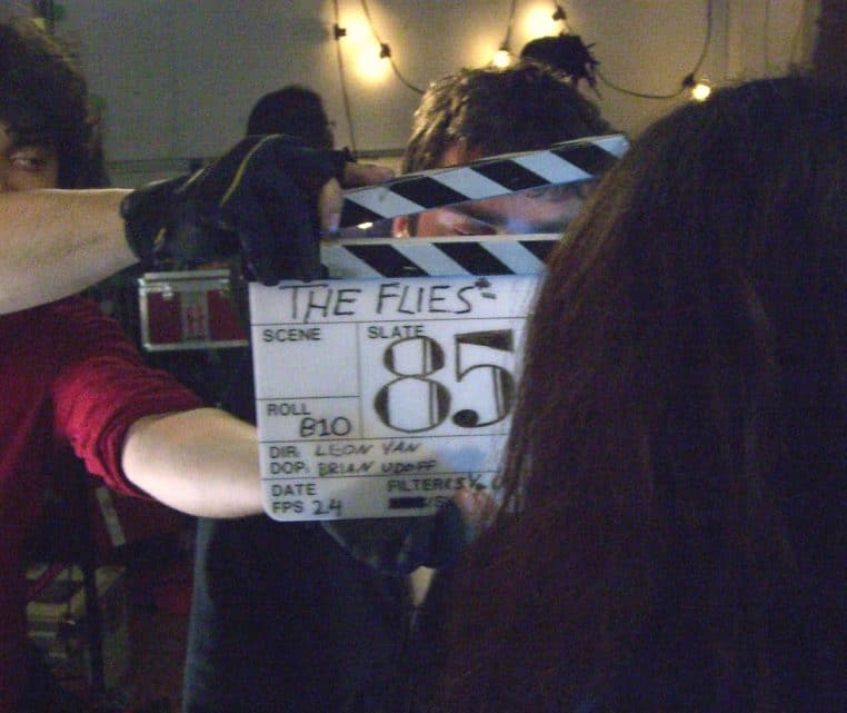 Clap de cinéma tenu par un assistant sur un plateau, lumière tamisée et équipe au second plan lors d’une prise.