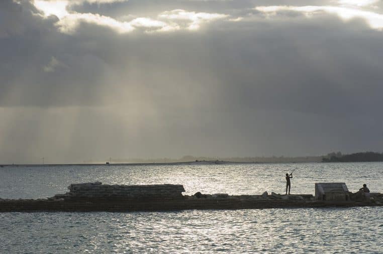 Berge sableuse bordée de cocotiers à Tarawa, houle légère sur lagon bleu et nuages élevés en fin d’après-midi.